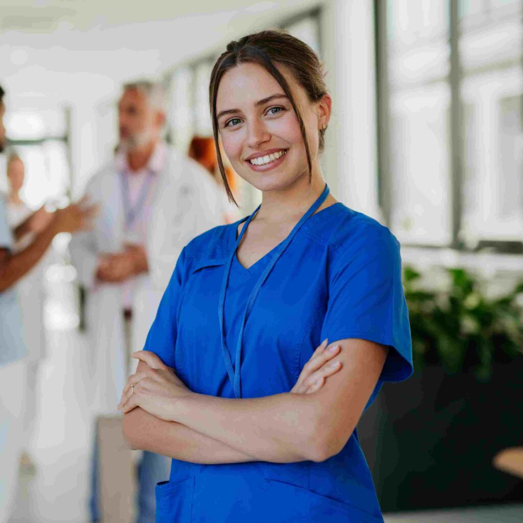 Junge Physiotherapeutin in blauer Arbeitskleidung steht lächelnd vor Kollegen in einer Klinik.