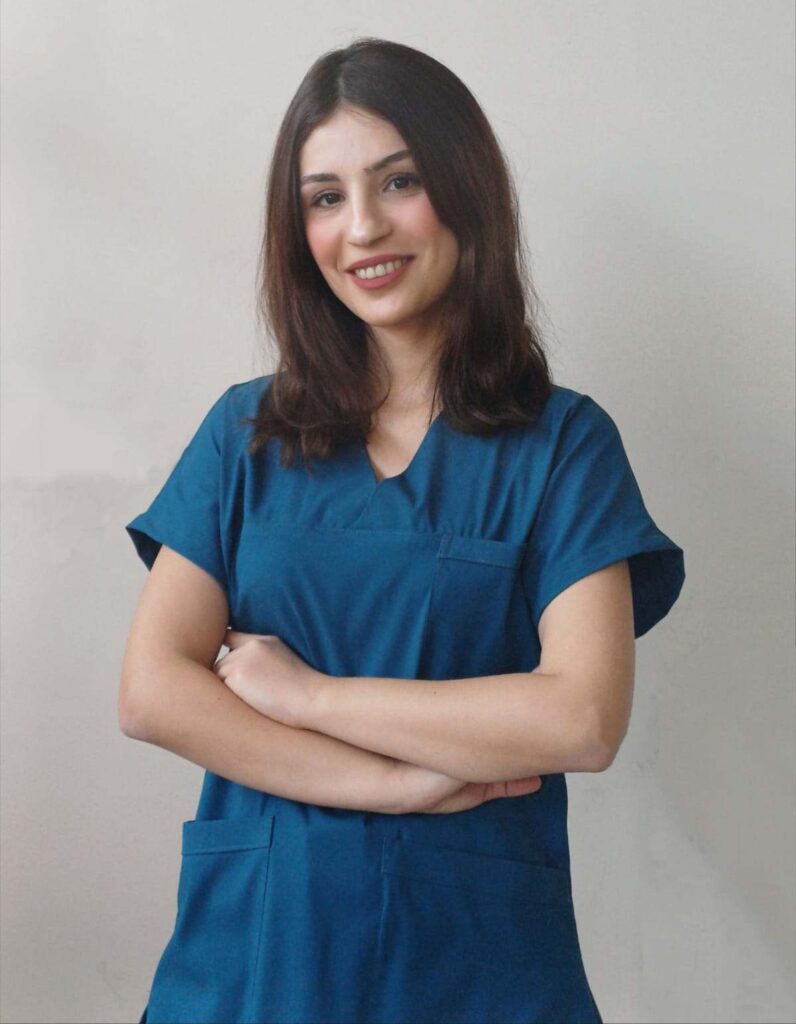 Physiotherapeutin Elif in blauer Berufskleidung, lächelnd mit verschränkten Armen, arbeitet als Physiotherapeutin in Anerkennung in Buttenwiesen.